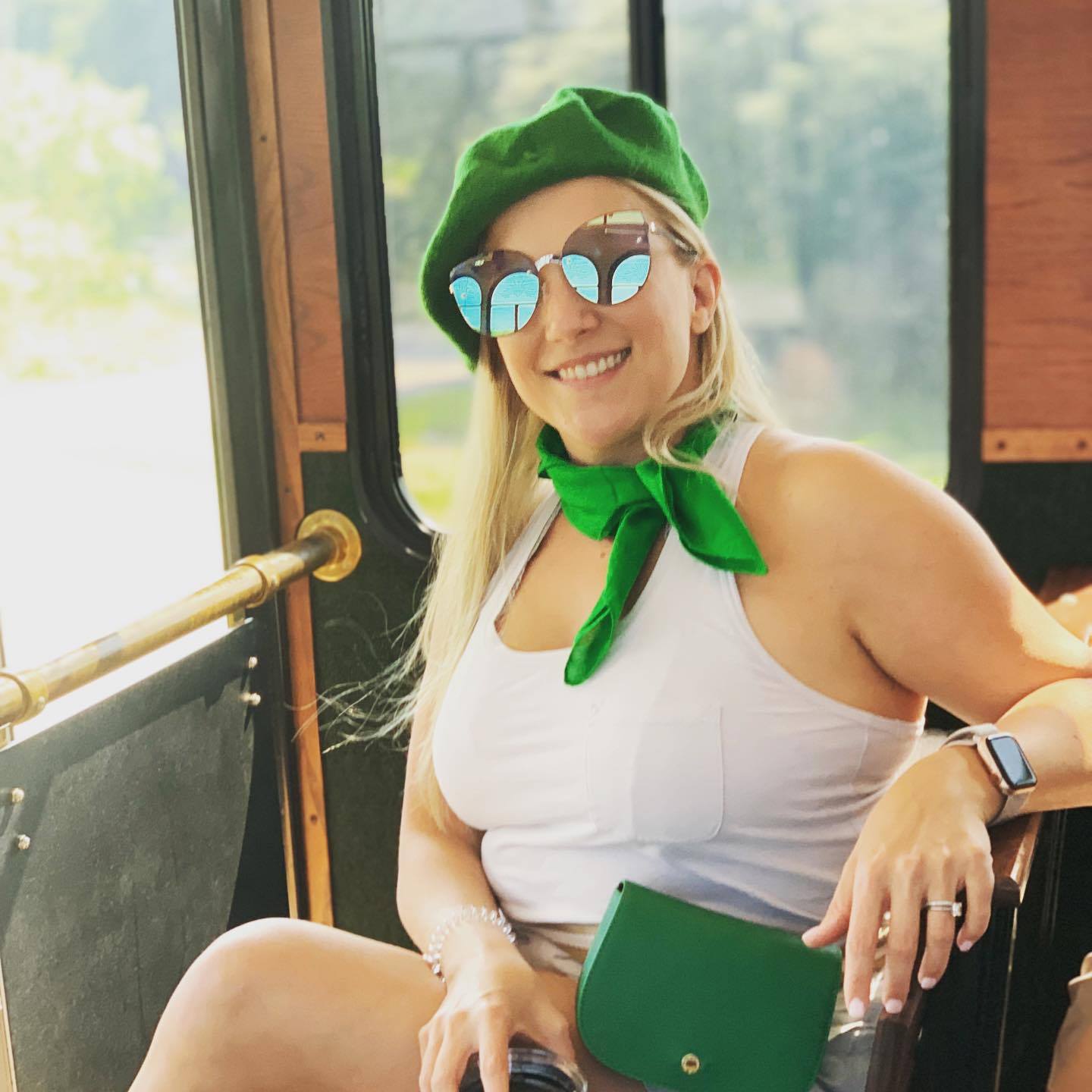 Ellen sitting inside a vintage trolley car, wearing a green beret, mirrored round sunglasses, and a green leather crossbody bag, grinning.